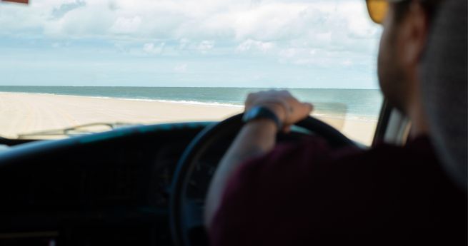 A person is driving a vehicle along a sandy beach with a view of the ocean in the distance. The photo is taken from inside the vehicle, focusing on the driver's arm holding the steering wheel.