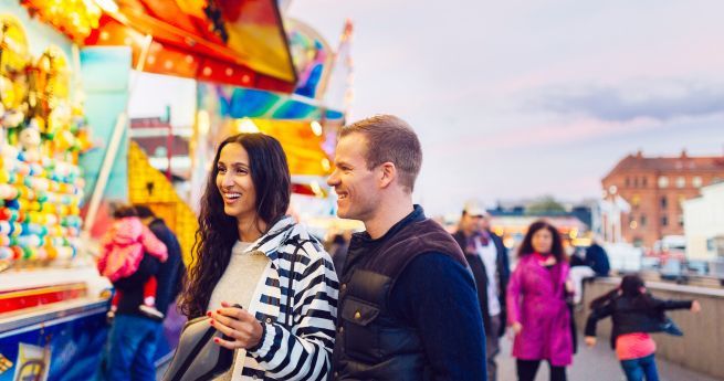 A smiling couple is enjoying a visit to a fair or carnival. The woman is wearing a striped jacket and holding a bag, while the man is dressed in a dark vest over a long-sleeve shirt. Bright, colorful game booths with prizes are visible in the background, along with other fairgoers.