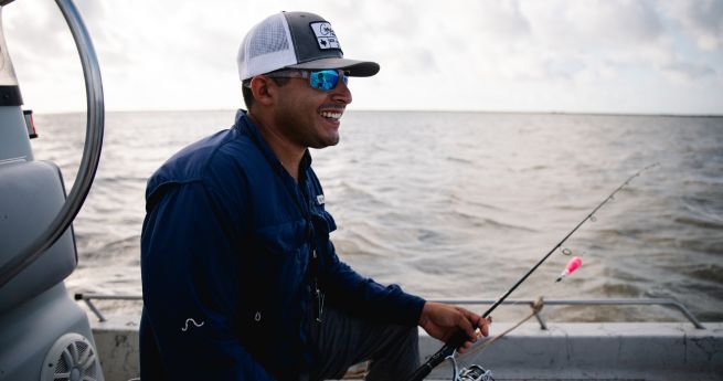 A man is smiling while fishing on a boat. He is wearing a blue jacket, a white and gray trucker hat, and reflective sunglasses. He is holding a fishing rod with a pink lure, and the background shows a large body of water under a cloudy sky.