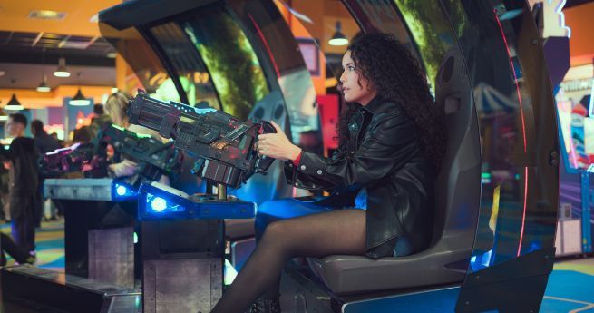 A woman with curly hair is sitting in an arcade game machine, intensely focused on playing. She is holding a large, futuristic-looking game controller or gun. The arcade is brightly lit with colorful lights and various arcade machines in the background. She is wearing a black leather jacket and sheer black tights.