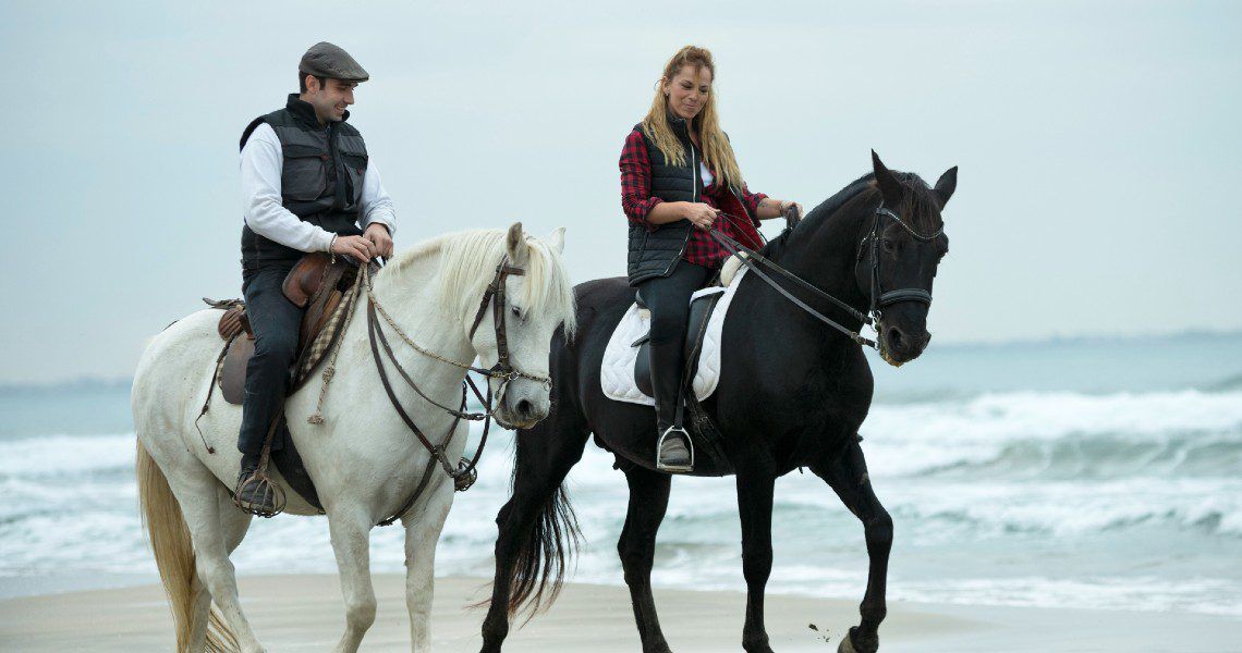 Two people are riding horses along a beach. The rider on the left is on a white horse and is dressed in a white shirt with a black vest and a cap. The rider on the right is on a black horse, wearing a red plaid shirt under a black vest.