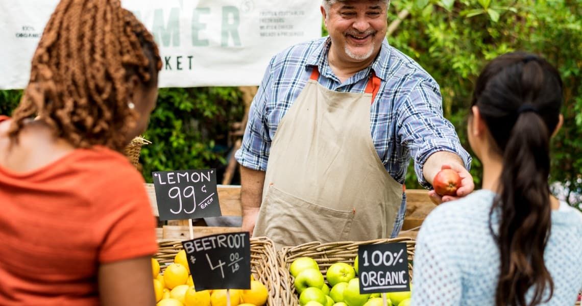 A friendly vendor at a farmers market is smiling and handing an apple to a customer. He is wearing a plaid shirt and a beige apron. The vendor stands behind a display of fresh produce, including lemons, beetroots, and green apples, with signs indicating prices and organic labels.