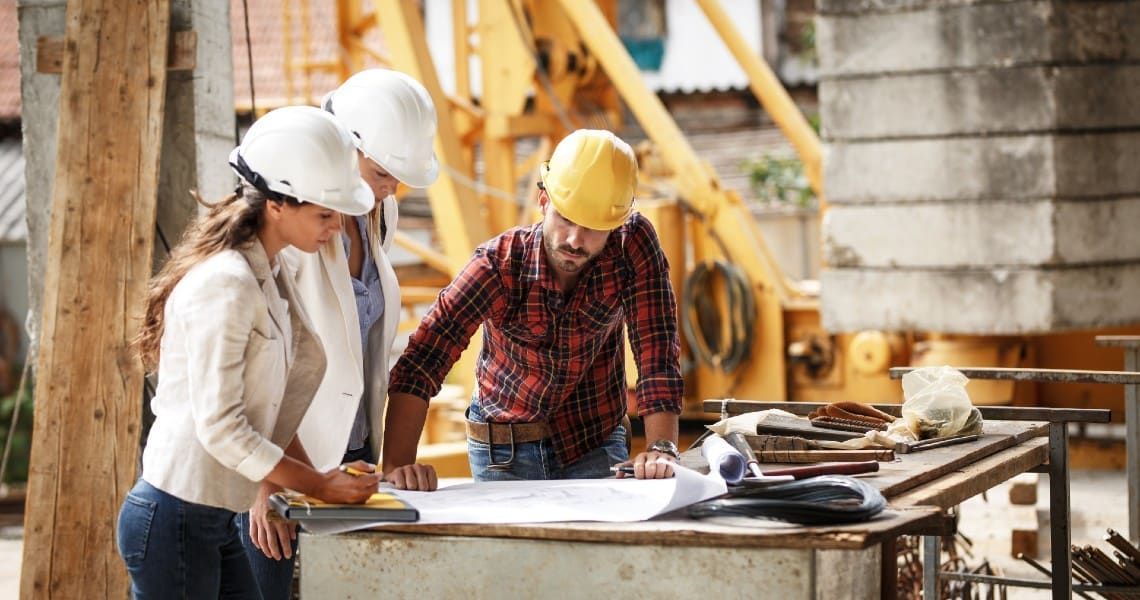 Three construction workers, two women and one man, are wearing white and yellow hard hats and reviewing blueprints on a construction site. They are standing around a table filled with various tools and materials, with construction equipment and structures in the background. The man, in a plaid shirt, is leaning over the plans, while the women, dressed in blazers, are attentively looking at the documents, discussing the project details.