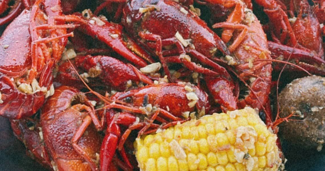 A close-up view of a delicious seafood boil featuring bright red crawfish seasoned with garlic and spices. The dish also includes yellow corn on the cob and a potato, all coated in flavorful seasoning.