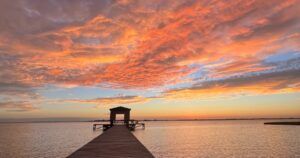 A stunning view of a wooden pier extending into calm waters under a vibrant, colorful sunset sky. The clouds are illuminated with shades of orange, pink, and purple, creating a dramatic and serene scene. At the end of the pier, there's a small structure silhouetted against the horizon, adding depth and focus to the composition.