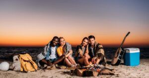 Four friends sitting on the beach around a campfire at sunset. They are wrapped in blankets, smiling and enjoying each other's company. The scene includes a cooler, a guitar, and a backpack, with the calm ocean in the background and a beautiful, vibrant sunset sky.