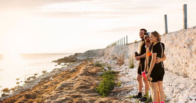 A group of three people standing near a coastal area, watching the sunset. They are dressed in athletic clothing and appear to be enjoying the view together.