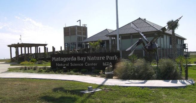 Entrance to Matagorda Bay Nature Park, featuring a modern building with a sloped roof and a sculpture of a large bird. The sign in front reads 'Matagorda Bay Nature Park Natural Science Center.' The surrounding area is landscaped with grass and native plants, and the sky is clear and bright.