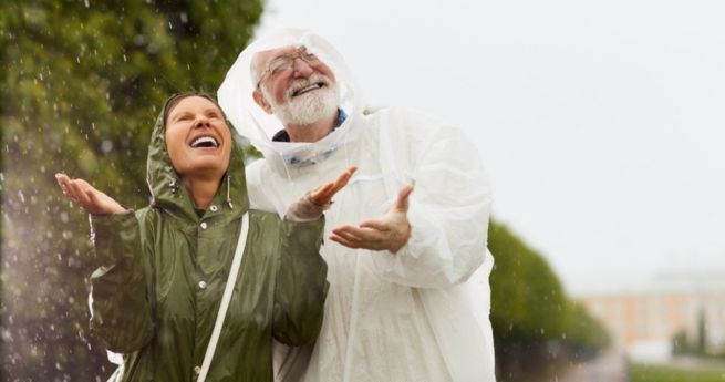 Two people enjoying the rain, dressed in raincoats. An elderly man wearing a white rain poncho and a younger woman in a green raincoat are smiling and looking up, with their hands raised to feel the rain.