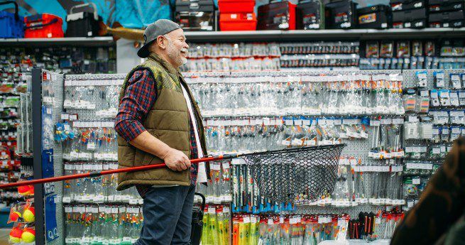 A man wearing a plaid shirt, a vest, and a backward cap is smiling and holding a large fishing net while shopping in a store filled with various fishing supplies. The shelves behind him are stocked with numerous fishing lures, hooks, and other equipment.