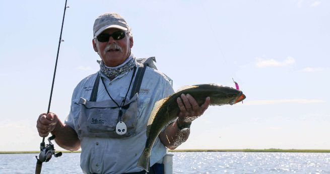 A man wearing a light-colored shirt, sunglasses, and a cap is standing on a boat while holding a fishing rod in one hand and a freshly caught fish in the other.