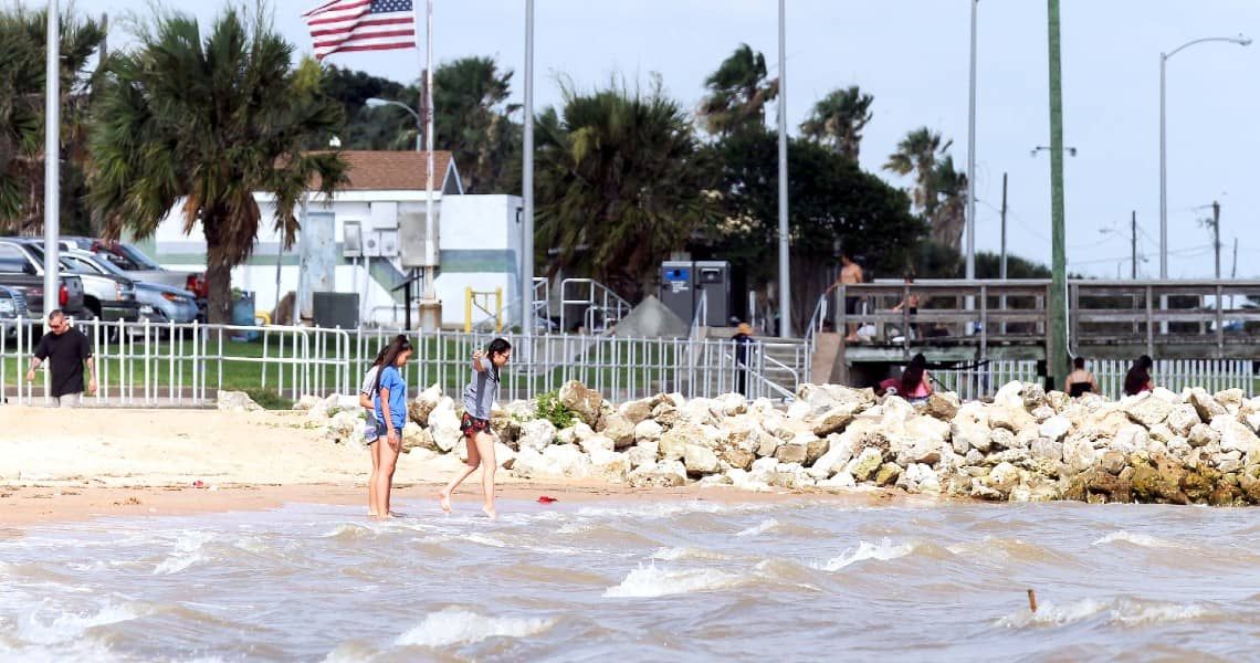 A lively scene at a beachside park with people enjoying the day near the water. Two young women walk along the shore, their feet in the gentle waves. Behind them, an American flag waves in the breeze, along with palm trees and a small building.