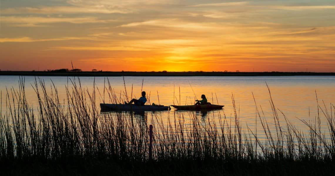 the best of palacios, tx-kayaking-4 A beautiful sunset scene featuring two kayakers on calm water. The sky is painted with warm hues of orange, yellow, and pink, reflecting on the water's surface.