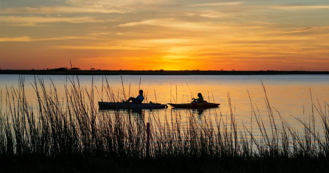 A beautiful sunset scene featuring two kayakers on calm water. The sky is painted with warm hues of orange, yellow, and pink, reflecting on the water's surface.