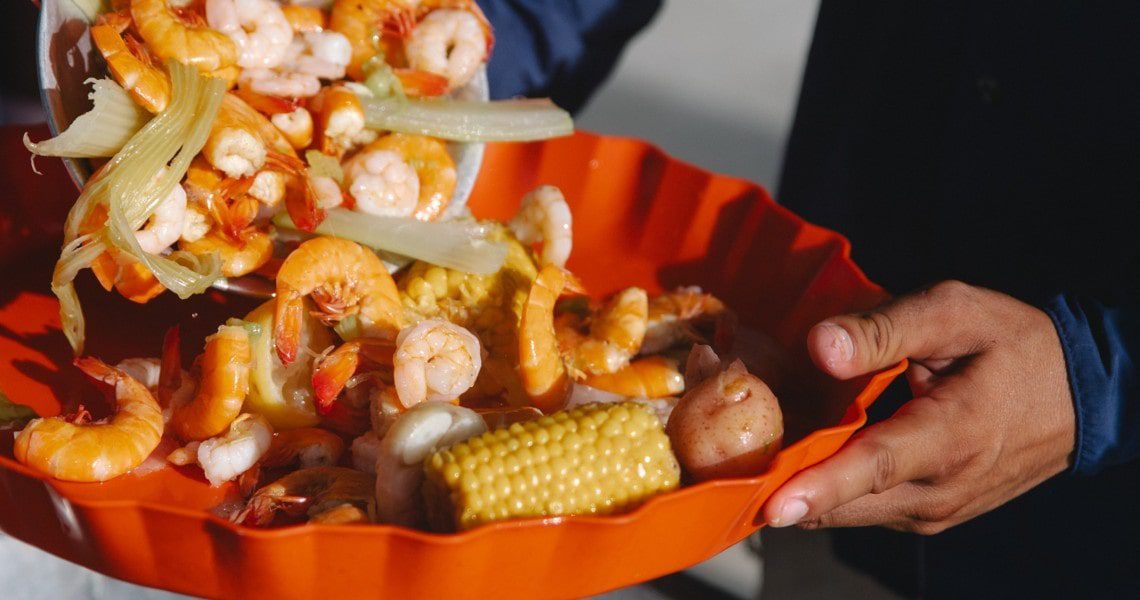 A vibrant and appetizing image of a seafood boil being served. The dish includes plump shrimp, corn on the cob, red potatoes, and celery, all arranged in a bright orange serving tray.