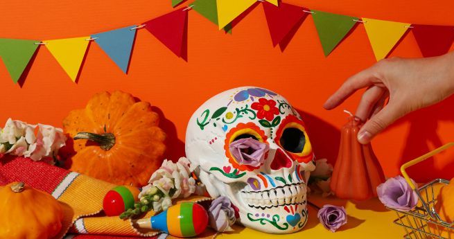 A colorful Día de los Muertos (Day of the Dead) scene featuring a decorated sugar skull with vibrant floral designs, surrounded by small pumpkins, flowers, and maracas. A hand is placing an orange candle next to the skull. The background is orange with multicolored triangular pennant banners hanging above, adding to the festive atmosphere.