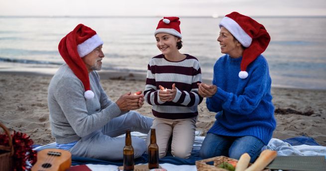 A joyful family Christmas scene at the beach, featuring an elderly man, a middle-aged woman, and a young girl, all wearing Santa hats. They are sitting on a blanket by the shore, smiling and holding lit candles. In the foreground, there are bottles and a guitar.