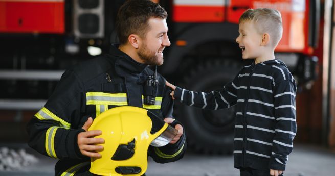 A firefighter in uniform is kneeling and smiling at a young boy, who is wearing a striped hoodie and smiling back. The firefighter is holding a yellow helmet, and they appear to be in a fire station with a fire truck in the background.