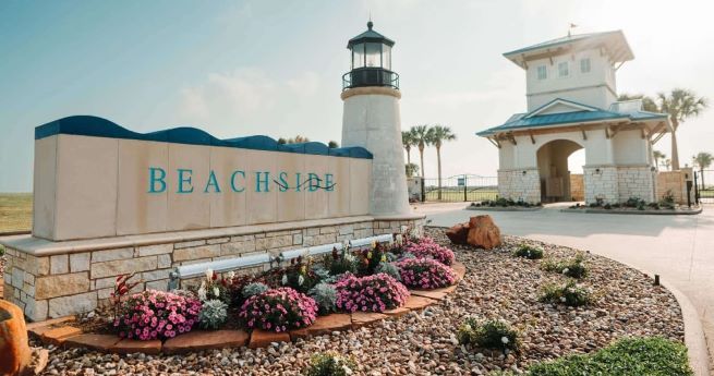 A welcoming entrance to a seaside community features a large stone sign with the word "BEACHSIDE" in teal letters. Beside the sign stands a lighthouse structure, adding to the coastal theme. The landscaping around the sign includes well-maintained flower beds with colorful blooms and rocks. In the background, there is a building with a blue roof and stone facade.