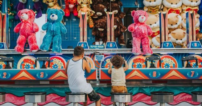 Two children seated on stools at a carnival game booth, aiming water guns at targets. The booth is decorated with large plush toys, including pink and blue teddy bears, as prizes.