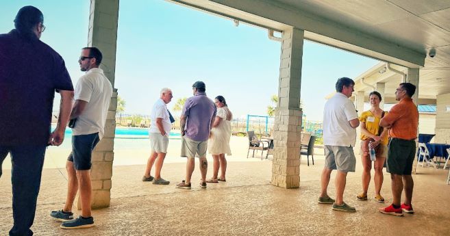 Group of people socializing at an outdoor event, under a covered patio area. The scene overlooks a swimming pool and a sunny day. The attendees are dressed in casual summer attire, engaging in conversation in small groups.