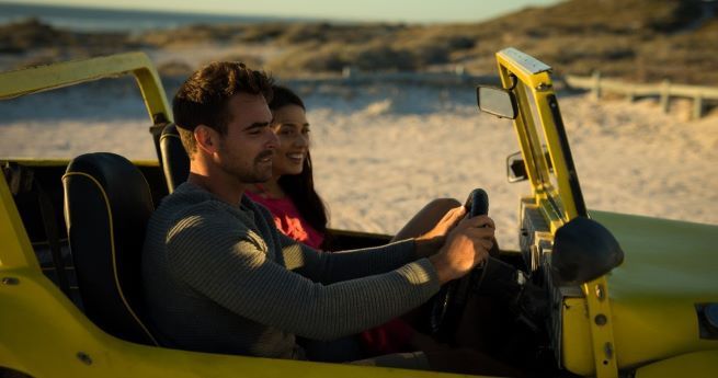 The image shows a couple enjoying a drive along a sandy beach in a bright yellow open-top vehicle.