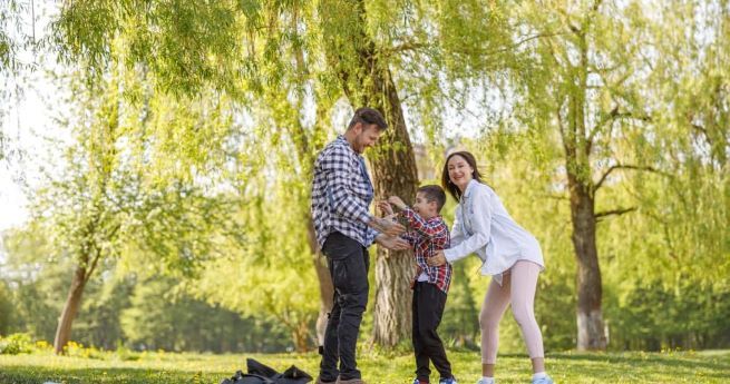 The image features a cheerful family enjoying time together in a lush, green park. The scene is centered on a father, mother, and young boy, all engaged in play.