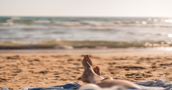 The image depicts a relaxed beach scene, focusing on a person lying on a sandy beach. The view is from the person's perspective, looking toward the ocean. Only the lower legs and feet of the person are visible, with toes pointing upwards. The beach is covered in soft, golden sand, and the waves gently lap at the shore in the background. The ocean is calm, with a few small waves, and the sun casts a warm, golden light over the scene.