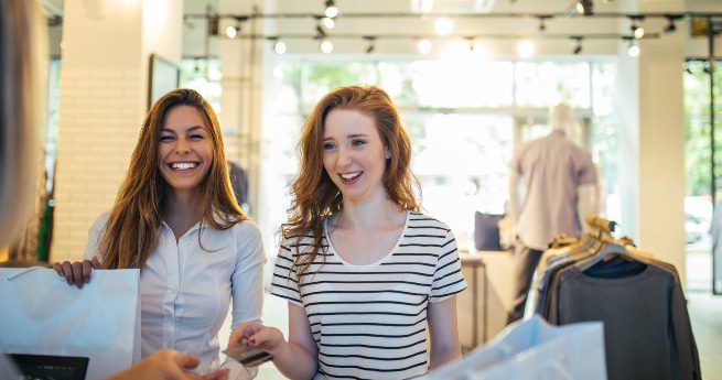 Two young women are enjoying a shopping experience in a bright, modern boutique. Both are smiling and appear to be in a good mood. The woman on the left has long brown hair and is wearing a light-colored blouse, while the woman on the right has red hair and is wearing a striped shirt. They are standing near the checkout counter, where one of them is handing over a credit card or payment to a cashier, out of frame. Shopping bags and racks of clothing are visible in the background.