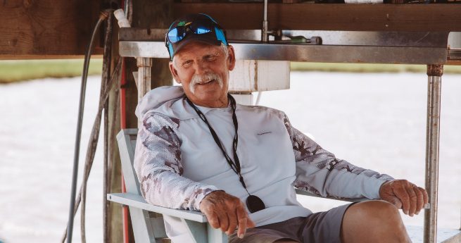 12 top things to do in port lavaca-fisherman-2 An elderly man is sitting comfortably in a chair outdoors under a wooden structure. He has a warm, relaxed smile, a white mustache, and is wearing a light-colored, long-sleeved shirt with a camouflage pattern on the sleeves.