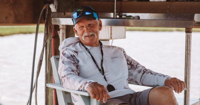 An elderly man is sitting comfortably in a chair outdoors under a wooden structure. He has a warm, relaxed smile, a white mustache, and is wearing a light-colored, long-sleeved shirt with a camouflage pattern on the sleeves.