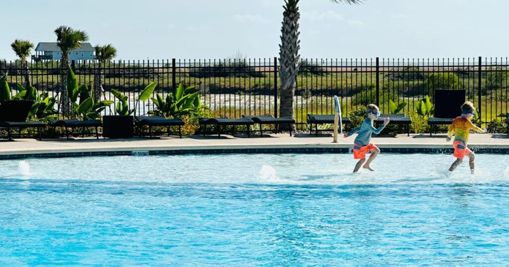 Two young children wearing colorful swim trunks and goggles are running and splashing in a shallow section of a swimming pool.