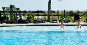 Two young children wearing colorful swim trunks and goggles are running and splashing in a shallow section of a swimming pool.