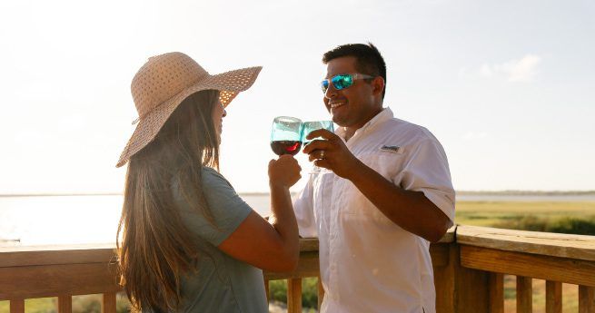 A man and a woman are enjoying a moment together on a wooden deck overlooking a scenic, sunlit landscape. The woman, with long brown hair, is wearing a wide-brimmed straw hat and a light-colored shirt, while the man is wearing a white short-sleeved shirt and blue reflective sunglasses. Both are smiling and holding up glasses of wine, toasting each other.