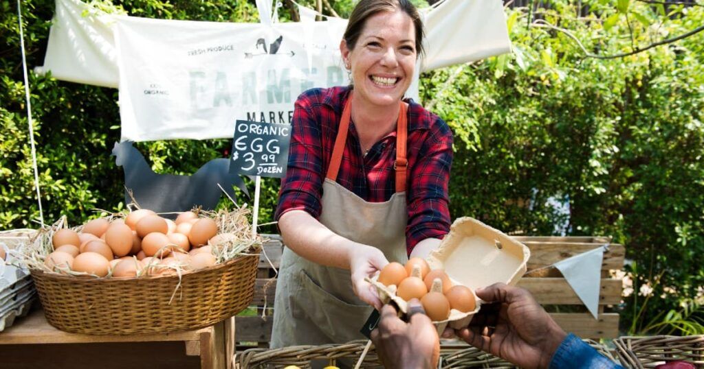 A smiling woman is selling eggs at a farmer's market. She is wearing a red plaid shirt with an apron and stands behind a table filled with baskets of eggs. The woman is handing a carton of eggs to a customer. Behind her, there is a sign that reads "Farmers Market" and another smaller sign advertising organic eggs for $3.99 per dozen. The market setting is outdoors, with greenery visible in the background.