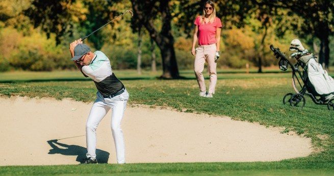 A golfer mid-swing, hitting the ball out of a sand bunker on a golf course. He is focused on his shot, wearing white pants, a light-colored shirt, and a cap. In the background, a woman in a pink polo shirt and light-colored pants watches the shot, standing near a golf trolley with a bag of clubs.