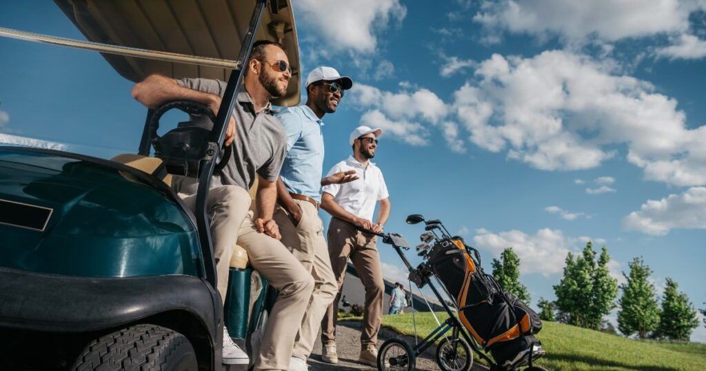 Three men enjoying a sunny day at the golf course. One man is seated in a golf cart, leaning on the side and gazing out at the course. The other two men stand beside the cart, chatting and smiling, with their golf bags on trolleys nearby. All are dressed in golf attire, including polo shirts, caps, and sunglasses.