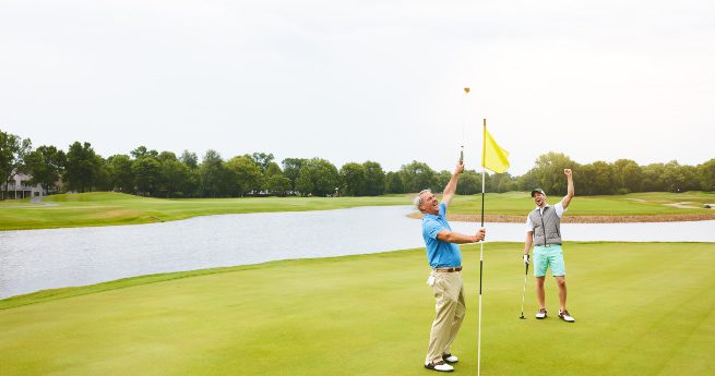 best bay city, texas, golf courses-two men celebrating -2 Two men celebrating on a golf course green beside a flagstick. One man is in the process of removing the flag from the hole, while the other raises his fist in excitement, indicating a successful shot or putt. Both are dressed in casual golf attire, with the backdrop featuring a serene lake, lush green grass, and a line of trees under an overcast sky.