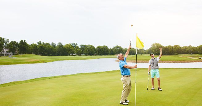 Two men celebrating on a golf course green beside a flagstick. One man is in the process of removing the flag from the hole, while the other raises his fist in excitement, indicating a successful shot or putt. Both are dressed in casual golf attire, with the backdrop featuring a serene lake, lush green grass, and a line of trees under an overcast sky.