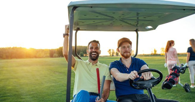 Two men sitting in a golf cart, smiling and enjoying a sunny day on the golf course. The man on the left is holding a golf club and has a cheerful expression, while the man on the right is at the wheel, looking content. The sun is setting in the background, casting a warm glow over the green landscape. In the distance, two other golfers are seen, one pushing a golf trolley.