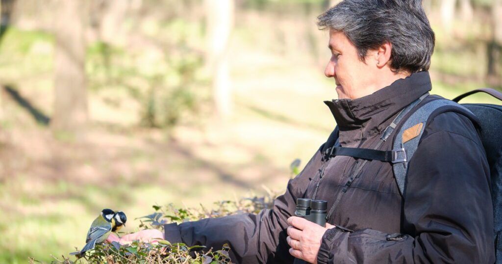 The image shows a woman outdoors, holding binoculars in one hand and gently extending the other hand towards a small bird perched on a bush in front of her.