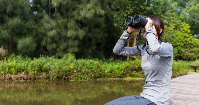 The image shows a woman sitting on a wooden boardwalk near a body of water, intently looking through a pair of binoculars.