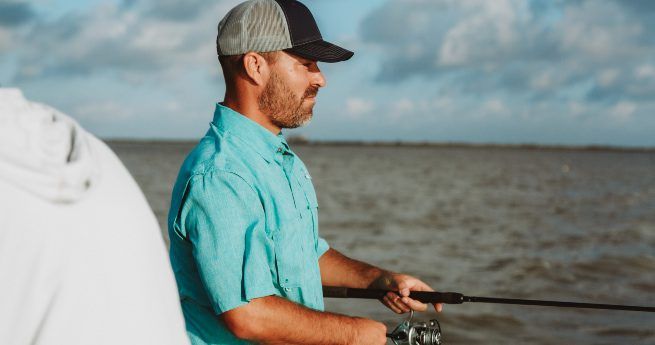 The image shows a man fishing, standing near the edge of a body of water. He is wearing a light blue short-sleeve shirt and a baseball cap with a dark front and mesh back. The man has a focused expression as he holds a fishing rod with both hands, his gaze directed toward the water.