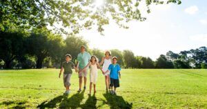 A happy family of five is walking hand-in-hand through a lush, green park under the shade of large trees.