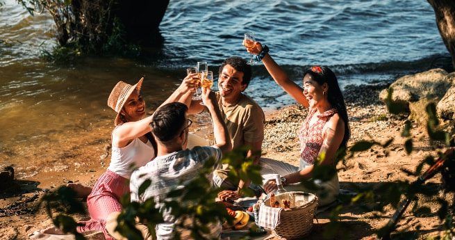 A group of four friends is enjoying a picnic by the river, sitting on the sandy shore with trees and water in the background. They are raising their glasses in a celebratory toast, smiling and laughing together. The picnic setup includes a woven basket and various snacks laid out on a blanket.