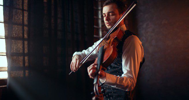 The image shows a man playing a violin in a dimly lit room. He is dressed in formal attire, wearing a vest over a long-sleeved shirt.