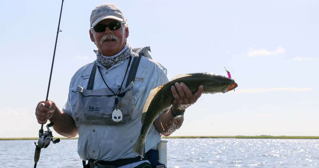 fantastic things to do in victoria, texas-man with fish-3 The image shows a fisherman proudly holding up a fish he has just caught. He is dressed in fishing gear, including a light-colored shirt, a cap, sunglasses, and a neck gaiter for sun protection. The fish is fairly large, with a shiny, sleek body, and the fisherman is holding it horizontally with one hand while holding a fishing rod in the other. The background features a calm body of water with a clear blue sky and a distant horizon.