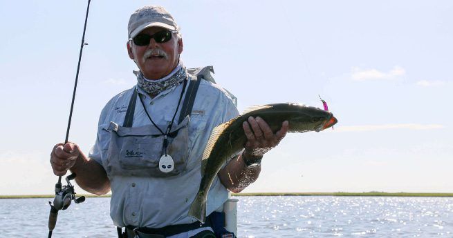 The image shows a fisherman proudly holding up a fish he has just caught. He is dressed in fishing gear, including a light-colored shirt, a cap, sunglasses, and a neck gaiter for sun protection. The fish is fairly large, with a shiny, sleek body, and the fisherman is holding it horizontally with one hand while holding a fishing rod in the other. The background features a calm body of water with a clear blue sky and a distant horizon.