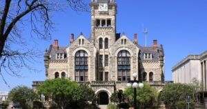 The image shows a historic courthouse with a clock tower situated in the center of its facade. The building is constructed from light-colored stone with intricate architectural details, including arched windows and decorative elements.