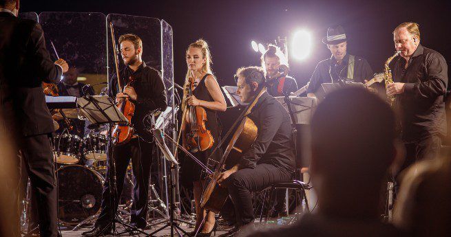 The image shows an orchestra performing on stage, featuring a mix of string and wind instrument musicians. The performers are dressed in black, with the string players holding violins, violas, and a cello, while one musician plays a saxophone.